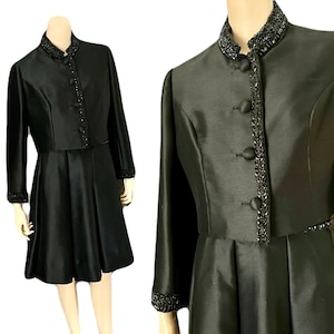 May include: A black two-piece formal dress set. The set includes a knee-length dress and a matching jacket. The jacket has a beaded collar, cuffs, and button details. The dress has a flared skirt. The fabric has a subtle sheen.