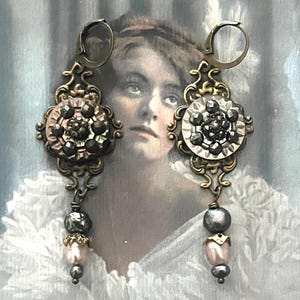 May include: A pair of ornate, vintage-style earrings. Each earring features a decorative, bronze-toned filigree design with a central cluster of dark, faceted stones. Below, a dangling arrangement of beads in gray and pink hues adds a touch of elegance.