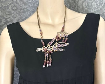 May include: A gold necklace with a purple and white crystal pendant in the shape of a branch. The pendant has a dangling chain with purple and white crystals.