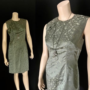 May include: A sleeveless, knee-length dress in a muted green colour with a subtle floral pattern. The dress has a round neckline and a fitted silhouette. A silver chain necklace with a pendant is worn over the dress.