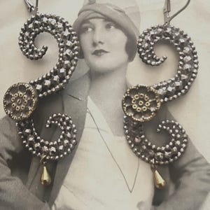 Puede incluir: Un par de pendientes plateados adornados con un diseño geométrico en espiral. Cada pendiente presenta un acento circular dorado y un pequeño colgante dorado en forma de lágrima. Los pendientes se muestran sobre una fotografía vintage en blanco y negro.