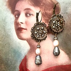 Può includere: Orecchini in stile antico dall'estetica vintage. Gli orecchini presentano un design circolare e ornato con un gruppo di piccoli abbellimenti argentati. Sotto, una combinazione di perle e una perla a forma di goccia grigio scuro pendono, creando un look unico ed elegante.