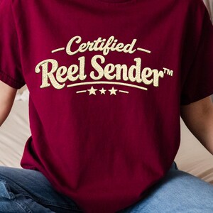May include: A maroon t-shirt with the text "Certified Reel Sender" in a gold glitter font. The shirt has a crew neck and short sleeves. The text is above three small stars.