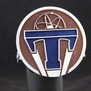 May include: A silver and brown circular metal emblem with a blue "T" in the centre. The "T" is surrounded by a white outline and a silver border. The background is brown with a stylised atom symbol.