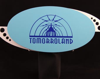 Può includere: Un cartello ovale azzurro con la scritta "TOMORROWLAND" in blu scuro. Il cartello presenta una grafica stilizzata di un edificio e archi. Il cartello è montato su una base nera e ha un accento argentato con ritagli circolari.