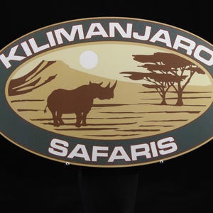 Può includere: Cartello ovale con una scena safari raffigurante un rinoceronte, alberi e montagne. Il cartello recita "KILIMANJARO SAFARIS" in lettere bianche su sfondo verde scuro e beige. Il design evoca avventura.