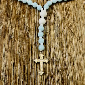 May include: A rosary with light blue and white beads. The rosary features a gold-colored cross pendant with a decorative design. The beads are strung together, creating a necklace-like appearance.