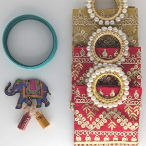 May include: A set of three decorative fabric pouches with gold embroidery and pearl trim. The pouches are stacked on top of each other, with the top pouch being a light gold color, the middle pouch being a red color, and the bottom pouch being a dark red color. The pouches are decorated with gold embroidery and pearl trim. A blue elephant figurine, two small glass bottles, and a teal bangle bracelet are also included in the image.