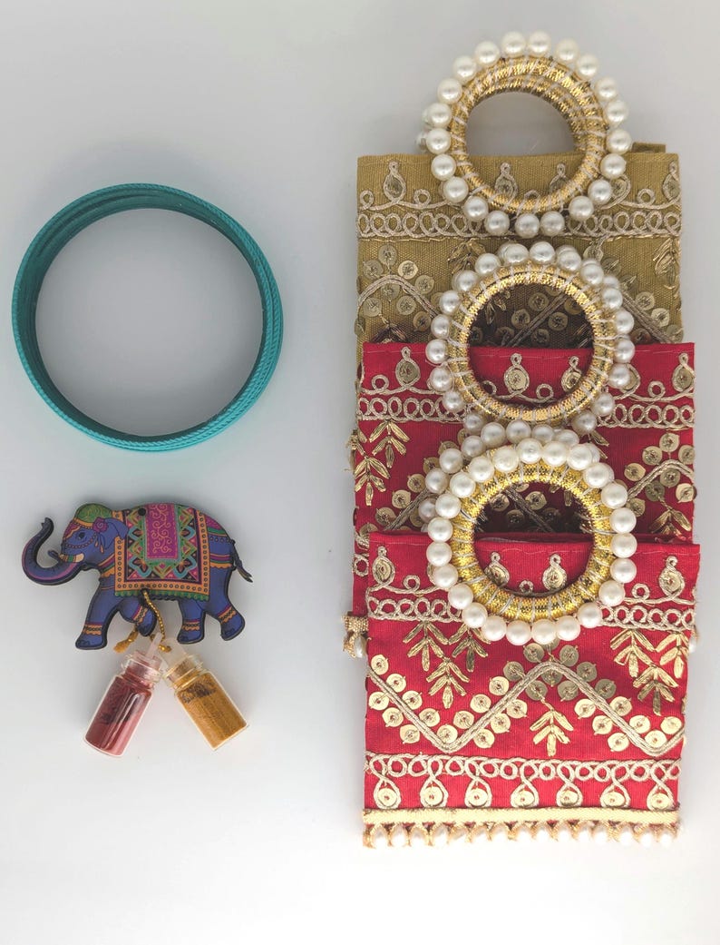 May include: A set of three decorative fabric pouches with gold embroidery and pearl trim. The pouches are stacked on top of each other, with the top pouch being a light gold color, the middle pouch being a red color, and the bottom pouch being a dark red color. The pouches are decorated with gold embroidery and pearl trim. A blue elephant figurine, two small glass bottles, and a teal bangle bracelet are also included in the image.