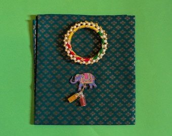 May include: A green fabric with a gold and white patterned design. A colorful bangle with red, yellow, and green stripes and white beads is on top of the fabric. A small wooden elephant figurine is below the bangle. A small bottle with a gold cap is hanging from the elephant's trunk.