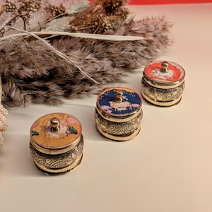 Könnte beinhalten: Drei kleine, runde, goldfarbene Behälter mit Deckeln. Jeder Behälter hat ein anderes farbenfrohes Design auf dem Deckel. Die Designs zeigen Tiere und Blumenmuster.