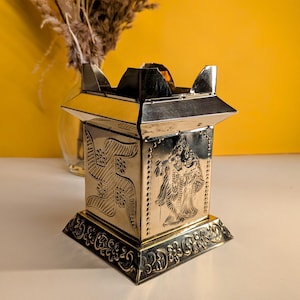 May include: A gold metal decorative stand with intricate carvings of a swastika and a deity. The stand has a square base and a rectangular top.