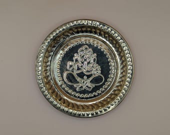 May include: A small, round, gold-coloured metal plate with a raised design of the Hindu deity Ganesha. The plate has a decorative border.