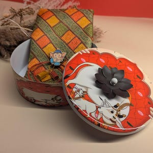 Op de afbeelding: Een ronde tinnen doos met een rood en wit ontwerp met een witte koe en een zwarte bloem. De deksel van de doos is open en onthult een groene en oranje gepatenteerde stof, een gouden en rode ketting en een klein blauw beeldje.