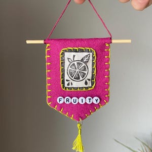 May include: A handmade magenta felt banner with yellow stitching and a wooden dowel. The banner features a black and white citrus fruit design and the word "FRUITY" spelled out in white letter beads. A yellow tassel hangs from the bottom.