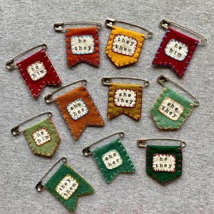 May include: A collection of handmade felt pronoun pins in various colors, including red, orange, yellow, and green. Each pin features embroidered text such as "he/him," "she/her," and "they/them," attached with a safety pin.
