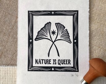 May include: A white rectangular paper print featuring a black ink block print of two ginkgo leaves, a small star, and the text NATURE IS QUEER, all enclosed in a decorative border. A wooden carving tool rests at the bottom right corner.
