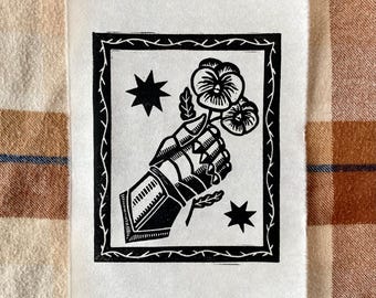 May include: Black and white linocut print featuring a gloved hand holding pansies, framed by a decorative border and stars. The text "Strength - Tenderness" is printed at the bottom. The print is on a white paper.