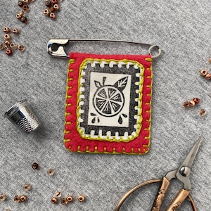 May include: A handmade red felt pin with a black and white citrus fruit design. The pin is attached to a silver safety pin. The image also includes a thimble, scissors, and beads, suggesting a crafting theme.
