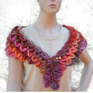May include: A crocheted shawl with a scalloped edge in shades of orange, pink, and purple. The shawl is worn by a person with a light brown shirt.