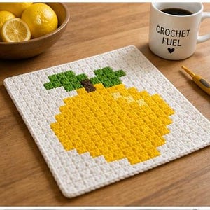 May include: A white crocheted square with a yellow lemon design and green leaves. A mug with the text "CROCHET FUEL" and a bowl of lemons are nearby. A crochet hook is also visible.