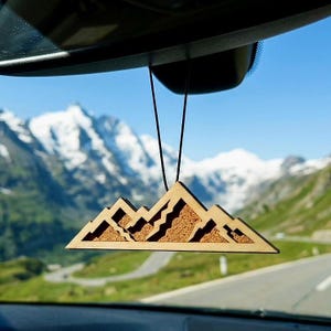 Pode incluir: Um ambientador de carro triangular de madeira com uma silhueta de cordilheira cortada a laser e uma incrustação de cortiça texturizada. O ornamento pende de um cordão preto fino, exibindo um design em camadas que imita picos e encostas.