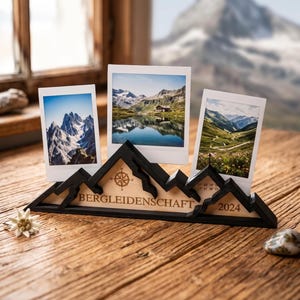 Peut inclure: Un support photo en forme de montagne en bois noir et clair, orné d'une icône de rose des vents, du texte BERGLEIDENSCHAFT et de l'année 2024. Le support maintient trois photographies rectangulaires verticales représentant des paysages de montagne.