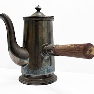 May include: Antique silver-toned coffee pot with a wooden handle. The pot has a long, curved spout and a lid with a small, pointed finial. The metal has a tarnished appearance, adding to its vintage charm.