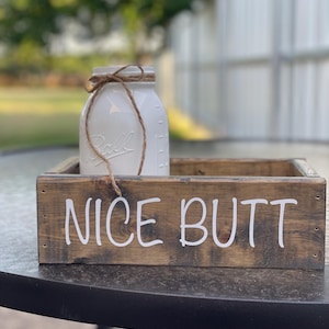 May include: A rustic wooden tray with the words "NICE BUTT" in white lettering. A white mason jar with a twine bow sits inside the tray. The jar has the word "Ball" printed on it.