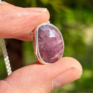 Może przedstawiać: Srebrny naszyjnik z wisiorkiem. Wisiorek ma wypolerowany, owalny, różowy kamień szlachetny, osadzony w srebrnej oprawie. Kamień szlachetny ma fasetowaną powierzchnię. Łańcuszek to delikatny, srebrny łańcuszek.