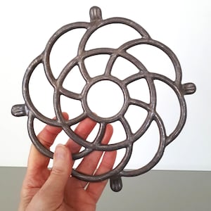 Peut inclure: Un dessous de plat en fonte en forme de fleur avec un centre circulaire et un motif en spirale. Le dessous de plat est gris foncé et a une finition rustique.