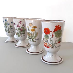 Puede incluir: Conjunto de cuatro tazas de cerámica blanca con diseños florales. Cada taza presenta una flor diferente con su nombre escrito debajo: "No me olvides", "Violeta", "Diente de león" y "Capuchina".