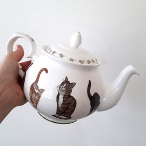 Op de afbeelding: Witte porseleinen theepot met een groene rand en een bloemenontwerp rond de deksel. De theepot heeft een grillig ontwerp met drie katten, een bruin-witte, een zwarte en een tabby.