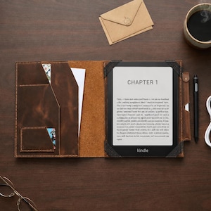 Può includere: Una custodia per e-reader in pelle marrone con tasche interne contenenti carta, un e-reader nero che mostra il testo Capitolo 1, uno stilo, un cavo di ricarica USB bianco, una busta marrone e occhiali con montatura dorata.