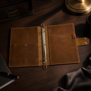May include: Open brown leather binder with a silver metal ring mechanism. The binder has pockets and a snap closure. A pen and notebook are visible on the dark wooden surface.