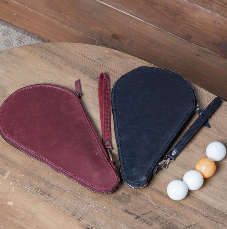 May include: Two leather paddle cases, one burgundy and one black, with zipper closures and straps. The cases are on a wooden surface with four white ping pong balls and one orange ping pong ball.