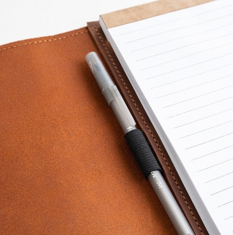 Op de afbeelding: Een bruin leren notitieboek met een gelinieerd notitieblok en een zilveren pen met een zwarte grip.