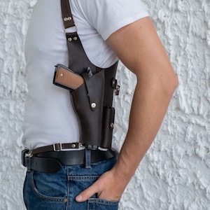 May include: A brown leather holster with a gun inside. The holster is worn over a white t-shirt and a black belt.