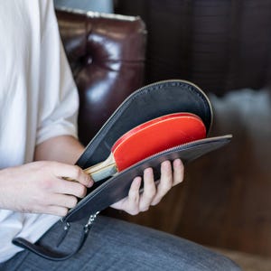 May include: A black leather carrying case with a zipper closure, open to reveal two red and black ping pong paddles.