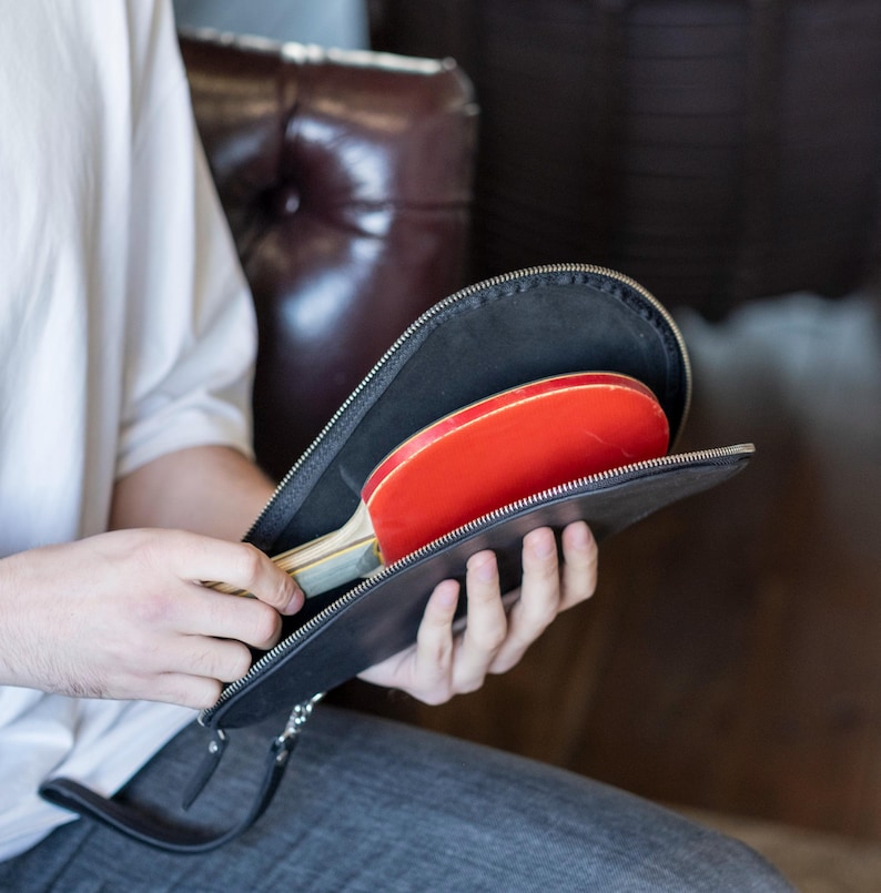 May include: A black leather carrying case with a zipper closure, open to reveal two red and black ping pong paddles.