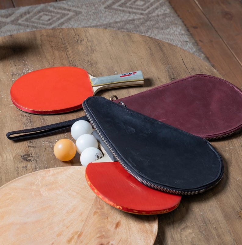 May include: Two red and black ping pong paddles with a black leather carrying case and four white ping pong balls on a wooden table.