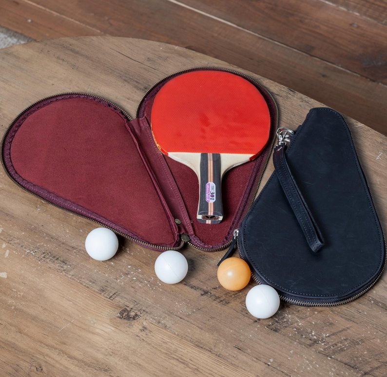 May include: A black leather carrying case with a zippered opening, holding a red and black ping pong paddle and three white ping pong balls. The case has a black leather strap.