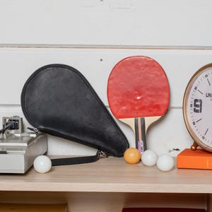 May include: A black leather ping pong paddle case, a red and black ping pong paddle, and three white ping pong balls on a wooden shelf. There is a clock with a white face and black numbers on the shelf behind the ping pong equipment.