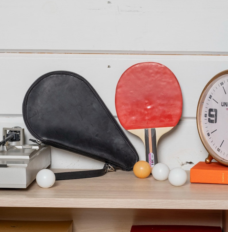 May include: A black leather ping pong paddle case, a red and black ping pong paddle, and three white ping pong balls on a wooden shelf. There is a clock with a white face and black numbers on the shelf behind the ping pong equipment.