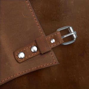 以下が含まれることがあります： 茶色の革のストラップのアップ。銀色のバックルとステッチが施されています。ストラップには銀色のリベットが3つ付いています。