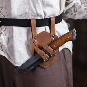 May include: A brown leather knife sheath with a black belt and a brown wooden handled knife. The sheath has two straps and is attached to the belt.