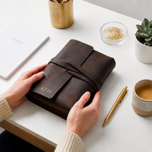 May include: A dark brown leather journal with a wrap-around strap closure and the embossed initials KFH on the front cover. The rectangular notebook features a smooth, matte finish and is held by two hands, showcasing its compact, portable design.