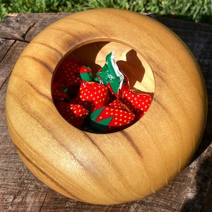 May include: A round wooden bowl with a light brown finish. The bowl has a circular opening in the center, filled with individually wrapped candies in red and green strawberry-themed packaging. The bowl is sitting on a wooden surface.