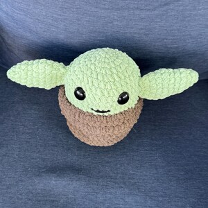 May include: A green crocheted plush toy with brown crocheted clothing and black eyes. The toy is shaped like a character from the Star Wars franchise.