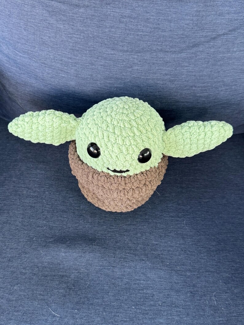 May include: A green crocheted plush toy with brown crocheted clothing and black eyes. The toy is shaped like a character from the Star Wars franchise.